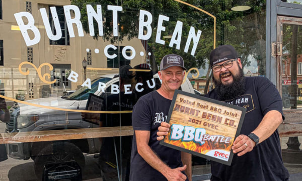Two men stand outside of Burnt Bean Co Barbecue holdinga best barbecue certificate, smiling.