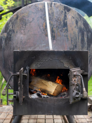 A close-up of a black metal wood-burning stove or smoker with a large cylindrical body, open front door showing burning wood and flames, and a white stripe down the center.