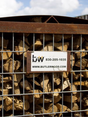 A metal wire mesh firewood cage filled with cut logs and branches, with a white sign reading 'bw 830-205-1035 WWW.BUTLERWOOD.