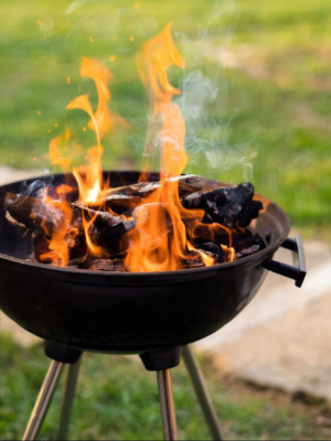 A black round charcoal grill with flames rising from burning coals, set outdoors on grass with a blurred green background.