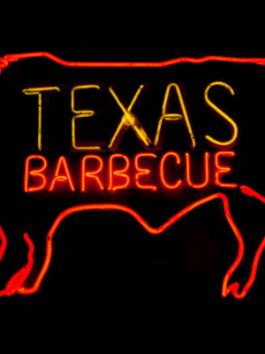 A neon sign featuring the words 'TEXAS BARBECUE' in glowing yellow and red letters, with red neon outlines of a pig  silhouette on a black background.