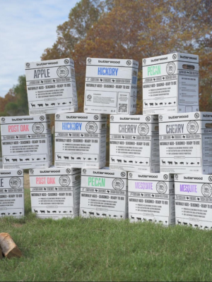 A stack of white wooden boxes printed with different flavor labels including Apple, Pecan, Cherry, and Pops Bark, arranged on a grassy lawn with trees in the background.