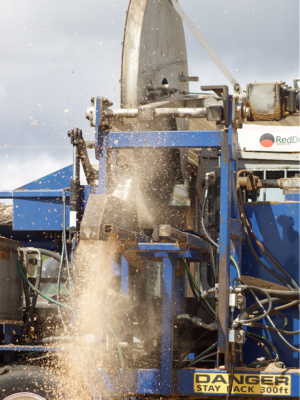 A close-up action shot of a large blue industrial machine in operation, with a large metal disc spinning and a stream of tan material or dust being blasted upward. A white cab is visible on the right with a RedDye logo, and a yellow warning label at the bottom reads 'DANGER STAY BACK 300ft'.