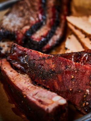 Close-up of glazed ribs with a dark charred exterior and seasoning, arranged on a baking sheet with sliced ham in the background.