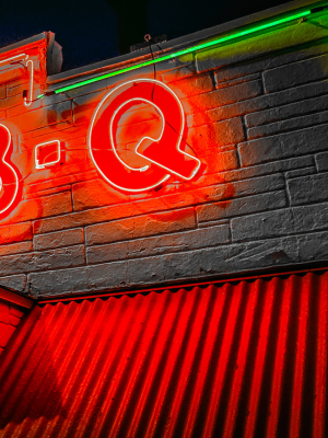 A glowing red neon sign showing large letters on a gray brick wall at night, with a bright green neon strip above and red light reflecting onto a corrugated metal surface below.