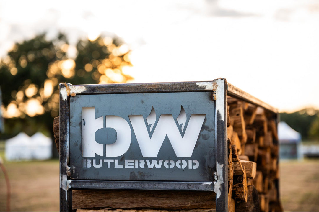 A rustic metal sign with the letters 'bw' and the text 'BUTLERWOOD' mounted on a stack of firewood outdoors at sunset. The sign is shown in a real environment with a blurred background of trees and tents.