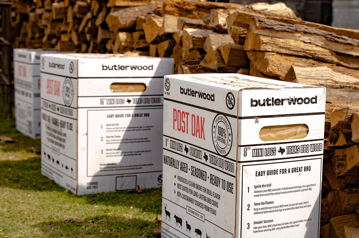 Several white Butlerwood wood storage boxes are stacked outdoors in front of a large pile of stacked firewood logs on grass.